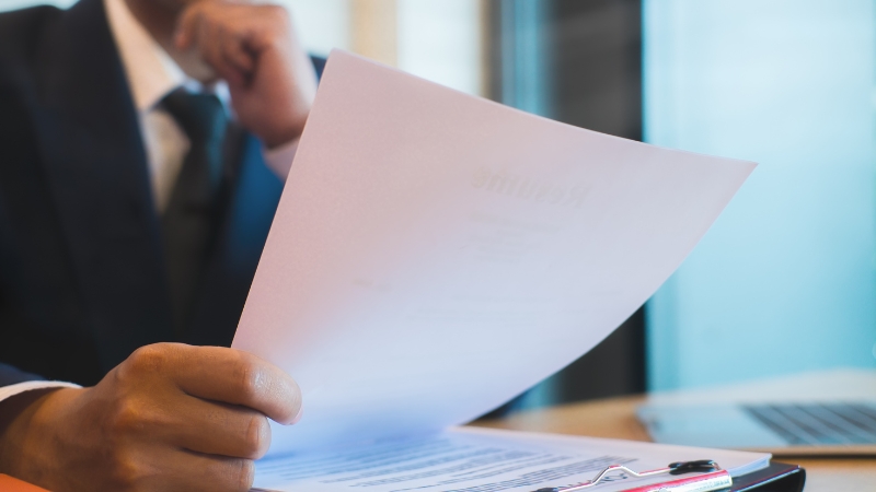 A person in a suit reviews a document at a desk with a clipboard and laptop nearby