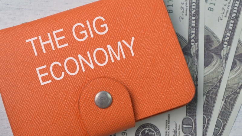An orange wallet labeled "The Gig Economy" lies on top of several hundred-dollar bills