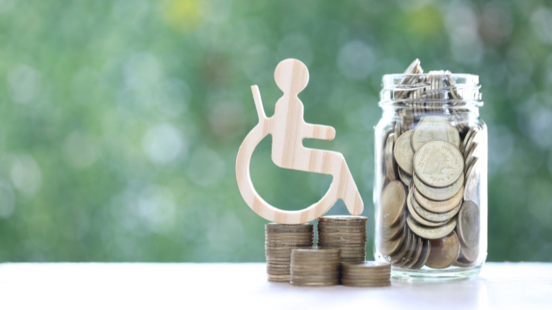 Wooden disability symbol beside stacked coins and a jar of money