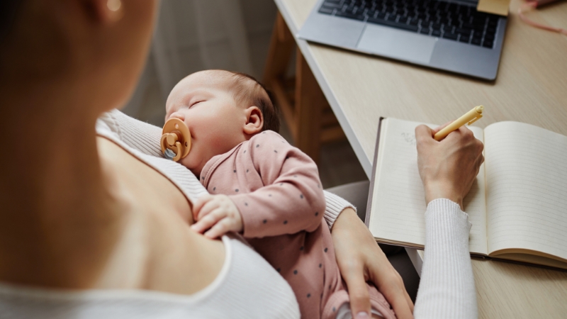 Mother holds a sleeping baby while managing tasks at home during maternity leave in the US